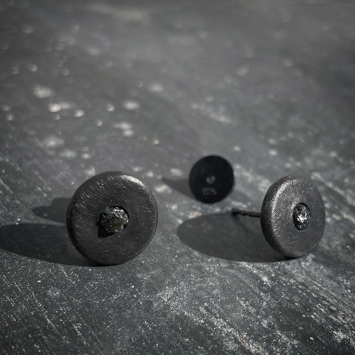 Black round ear studs with black raw diamond on a textured gray surface
